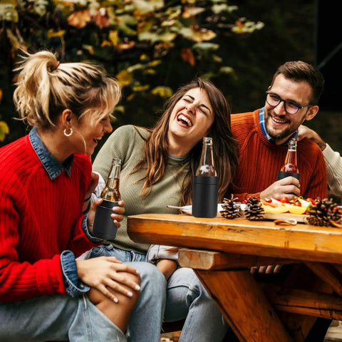 Group of friends enjoying drinks outdoors using Stainless Steel 12oz Vacuum Insulated Can And Bottle Cooler Keeps Ya Drinks Ice Cold With Double Wall Construction Copper Plated Chiller Metal Cooler For Cans And Bottles Black Finish on a picnic table in a forest setting.