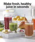 Juicer machine on kitchen counter with four glasses of colorful juice (red, green, orange) and fruit in background.
