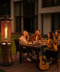 Gas Patio Heater With Wheels, Floor Standing Outdoor Heater With Round Dancing Flame And Glass Tube, Quick No Tool Setup Under Five Minutes, 20 Lb Propane Tank Compatible For Balcony Veranda And BBQ Party next to dining table on a patio.