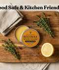 Real Deal Natural Beeswax Furniture Polish To Restore Dry Wood And Boost Natural Grain With Food Safe Olive Oil Blend For A Smooth Clean Finish That Gives Your Favorite Wooden Pieces Brand New Life In 3.5 Fl Oz Jar on a wooden cutting board with herbs and lemon.