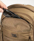 Person placing sunglasses into MOLLE Compatible Diaper Backpack for Dads With Built-In Changing Mat and Stroller Straps, 600D Coyote Brown.
