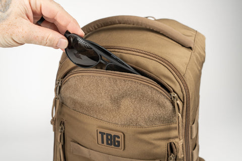 Person placing sunglasses into MOLLE Compatible Diaper Backpack for Dads With Built-In Changing Mat and Stroller Straps, 600D Coyote Brown.