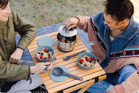 Two people using Wildfare Go 14 Piece Stainless Steel Camp Kitchen Set with 1.65 Quart Saucepan on wooden camping table