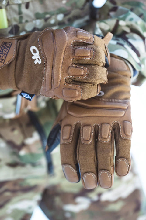 Person wearing brown tactical gloves with reinforced knuckles, set against a camouflage jacket, outdoor setting.