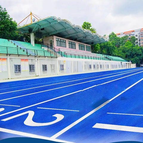 Blue athletics track with lanes marked, adjacent to grandstand, trees, and buildings.