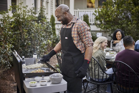 Man cooking on a griddle using the Griddle Essential 5 Piece Tool Set with spatula, scraper, and squeeze bottles, outdoor gathering