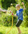 Garden hose, yellow with red spray nozzle, being used by a smiling child in a sunny backyard