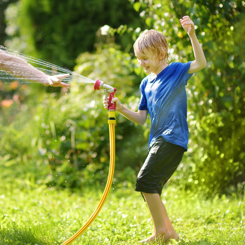 Garden hose, yellow with red spray nozzle, being used by a smiling child in a sunny backyard