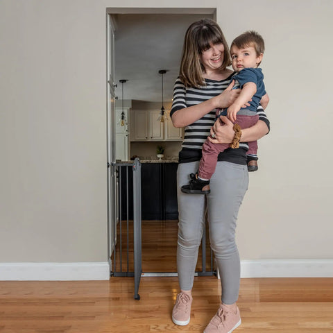 Woman holding child next to the Extra Tall Decor Safety Gate For Kids And Pets In Gray Fits Openings From 28 To 38.25 Inches Wide And 36 Inches High Perfect For Doorways Stairways And Between Rooms No Fuss Install With Extensions And Stair Kit in hallway.