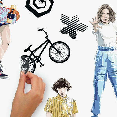 Hand applying Stranger Things Peel And Stick Wall Decals Set With 17 Pieces Removable And Repositionable Without Residue For Any Smooth Surface Perfect For Renters And Homeowners Seeking Retro Sci Fi Style And Easy Decor Makeover featuring retro sci-fi characters and symbols.