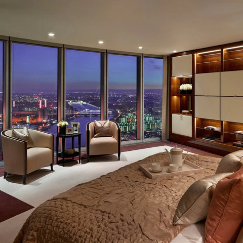 Luxury bedroom with city skyline view, featuring armchairs, side table, and plush bedding. Soft ambient lighting.