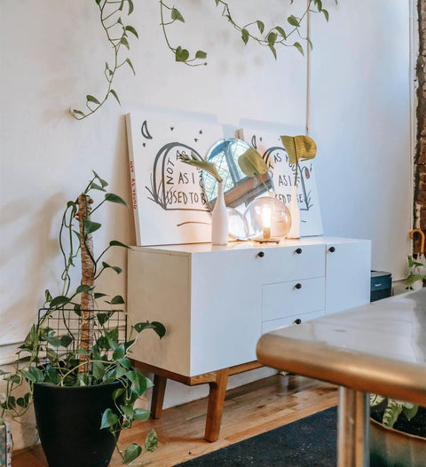 Modern white cabinet surrounded by green plants, featuring decor and lighting on top, minimal home interior design.