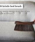 Soft bristle bed brush on gray couch, designed for cleaning fine debris, wood handle, narrow for crevices.