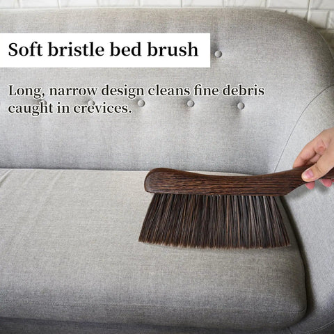 Soft bristle bed brush on gray couch, designed for cleaning fine debris, wood handle, narrow for crevices.