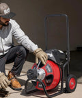 Man using Auto Feed Drain Cleaner On Wheels With 100 Ft Cable For 2 To 6 Inch Pipes, Portable Sewer Snake Auger outdoors.