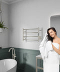 Woman in bathroom with Glow 4 Bar Wall Mounted Towel Warmer in Brushed Nickel, 40W stainless steel heated towel rack above tub.