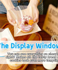 Person holding a 100 Pack 7.1 By 7.5 Inch Cookie Bags With Clear Window Sealing Stickers For Easy Packaging Grease Resistant Brown Paper Bakery Bags For Cookies Bread And Pastries showcasing a donut.