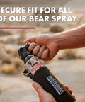 Person holding Frontiersman Max Bear Spray And Mountain Lion Deterrent With 40 Foot Range For Outdoor Folks Max Strength 2.0 Percent Major Capsaicinoids EPA Registered Self Defense For Hiking Camping Hunting Fishing And Biking 7.9 Oz with secure fit sleeve.