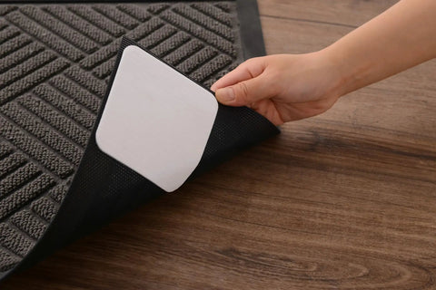 Hand applying Rug Gripper Set Of 4 Non Slip Corner Pads For Hardwood Floors And Tile on a rug edge, showing adhesive side on wood floor.