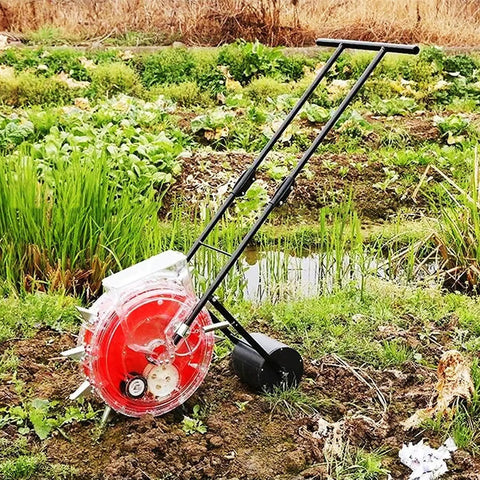 Manual Seed Planter And Fertilizer Applicator For Garden on grass, showing red roller seeder and iron handles in a field setting.