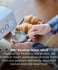 Person using Talos Semi Automatic Grind And Brew Espresso Machine With Built In Conical Burr Grinder 20 Bar Italian Pump Real Time Pressure Gauge Stainless Steel Lunar Finish For Barista Level Flavor At Home to froth milk with a steaming wand, with croissants in the background.