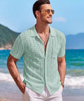 Man wearing Men's Knit Short Sleeve Button Down Shirt Vintage Polo Beach Top in Ice Green, standing on beach with mountains.