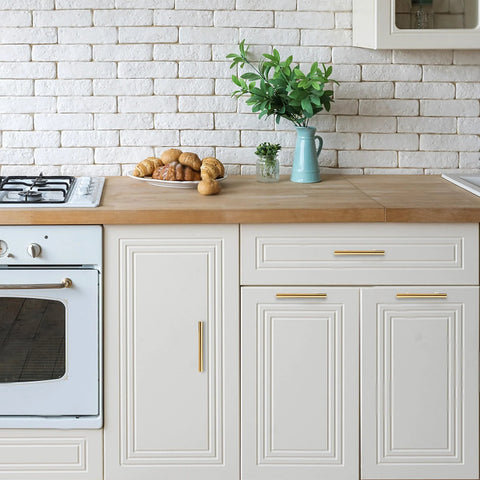 Modern kitchen with white cabinets and gold handles, showcasing countertop with bread and a plant on a wooden surface.