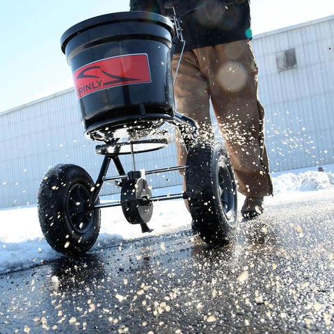 50 Lb Capacity Push Broadcast Spreader For Lawns And Gardens in use on snowy pavement, showing durable wheels and easy push handle.