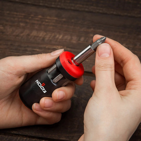 Hand holding Stubby Ratcheting Screwdriver Kit With 14-In-1 Mini Bit Set Including Flathead, Hex, Torx, Square, And Pozidriv Tips Compact Black And Red Dope Handy Tool For Quick Fixes And Electronics Projects on wooden background