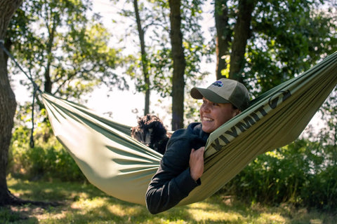 Person relaxing in a green camping hammock with dog in woods, enjoying outdoor adventures