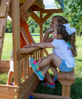 Children playing at snack bar window of Beachfront Cedar Wood Swing Set With Wave Slide Elevated Deck Canopy Monkey Bars Rock Wall Ladder Shaded Picnic Area Snack Bar Window Belt Swings Trapeze Sandbox Playset For Kids.