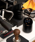 Subscale Digital Dosing Cup With LED Display Sleek Black Scale For Espresso Brewing next to espresso machine and tamper