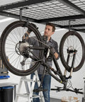 Man installing bike on Four Pack Garage Ceiling Bike Hooks Overhead Mount For Hanging Bicycles And Tools Heavy Duty 33 Lb Capacity 3x6 Inch L Shaped Hooks