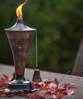 Oahu Tiki Style Outdoor Torch with brushed bronze finish, lit with flame, on deck surrounded by autumn leaves.