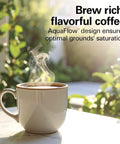 Cup of steaming coffee on a table showcasing AquaFlow design for rich flavor, plants in the background.