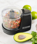 Black mini electric food chopper with 2 cup capacity, one touch operation, filled with chopped vegetables, next to avocado and cilantro.