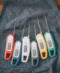 Array of ultra fast digital instant read meat thermometers with 3-4 second readouts, foldable probes on denim fabric.
