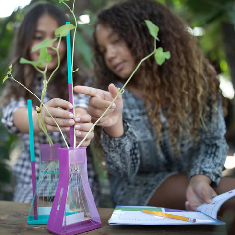 Transform Kids Into Junior Botanists With This Hella Awesome Stem Grow Kit Featuring Zero Hassle Indoor Soil Free Planting Plus Glass Flasks And Discovery Logs. Two girls examine plants in glass flasks.