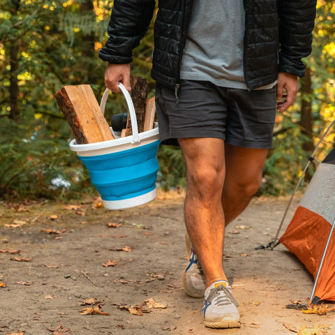 Person carrying a blue Collapsible 2.6-Gallon Camping Bucket with firewood by a tent, ideal for dishwashing, cleaning, and storage.