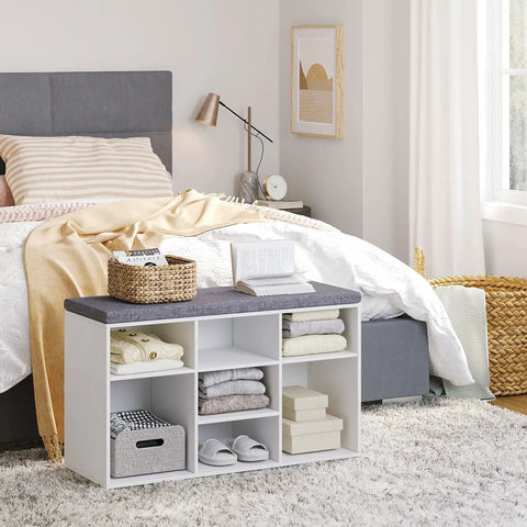 White cushioned shoe storage bench with 9 cubbies and adjustable shelves, placed in a bedroom next to a bed.