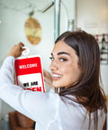 Person holding red and white office door open sign with suction cup, smiling indoors at a storefront.