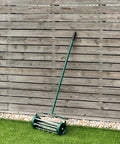 Rolling Lawn Aerator With 3 Piece Long Steel Handle And Fender 18 Inch Rotary Push Tine Spike Tool next to wooden fence