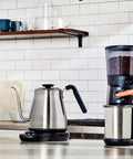 Gooseneck Electric Kettle For Pour Over Coffee And Tea With Dialed In Temperature In One Degree Increments From 104 To 212 Fahrenheit And 40 To 100 Celsius Built In Brew Timer Stainless Steel 1 Liter on a kitchen counter next to a coffee grinder.