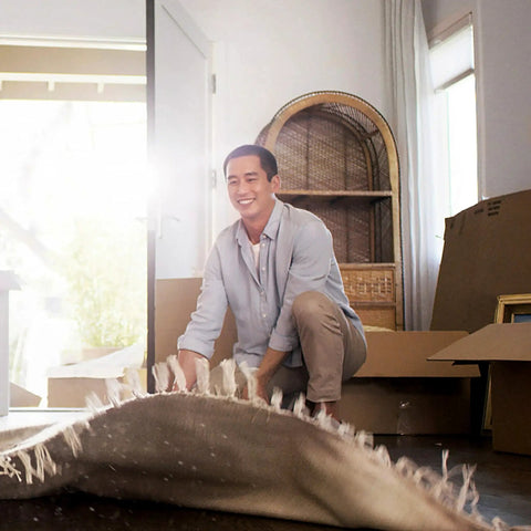 Smiling person kneeling in room with boxes and rolled up rug, bright natural light from window