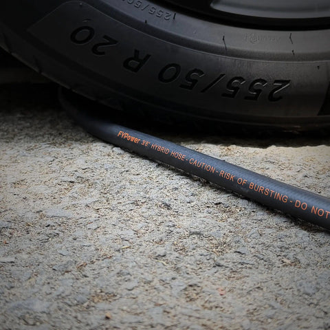 No Fuss Lightweight Hybrid Air Compressor Hose 3/8 In X 10 Ft With 1/4 Inch NPT Couplers Rated To 300 PSI beside a tire on gravel.