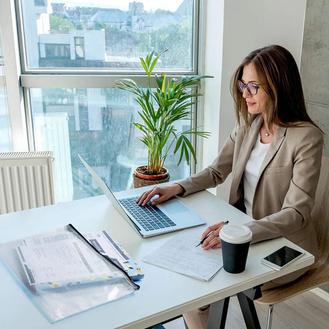 Woman working at a desk with laptop, coffee cup, and 12 Pack Waterproof Zipper File Folders with Tear-Resistant PVC Pouches