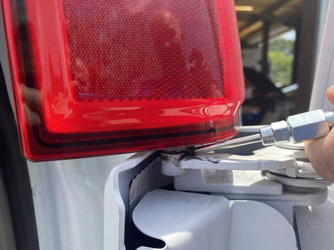 Grease gun needle nozzle, metal with steel tip, attached to hydraulic fitting beside red tail light on white vehicle