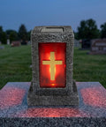 Red illuminated cross on stone memorial lantern in cemetery during dusk.