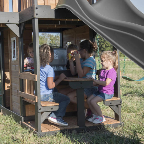 Children playing in cedar wood swing set with wave slide and elevated clubhouse play kitchen and picnic area.
