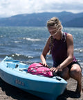 Floating Waterproof Dry Bag Backpack 10L Pink For Kayaking Rafting Boating And Outdoor Adventures next to a blue kayak on the shore
